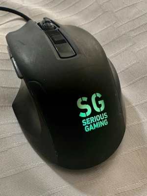 SG Serious Gaming USB Gaming Mouse - Ergonomisk gamingmus från Serious Gaming med USB-anslutning. Utrustad med flera programmerbara knappar och belyst SG-logotyp. Passar perfekt för spelentusiaster. Viss yttre slitage syns, men fungerar som den ska.