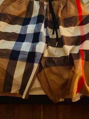 Burberry - Säljer ett par snygga Burberry shorts med klassiskt rutigt mönster i beige, brun, svart, vit och röd. De har bekväm snörning i midjan och är perfekta för dig som gillar en chill och trendig stil. Passar bra till sommaren och har en loose fit.