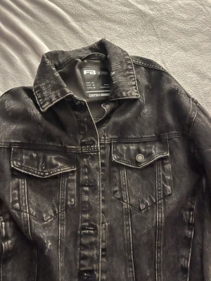 Svart jeansjacka från FB Sister - Cool svart jeansjacka från FB Sister med slitna detaljer och klassisk krage. Jackan har två bröstfickor med lock och knappar framtill. Materialet är denim och jackan har en snygg tvättad look som ger den extra edge.
