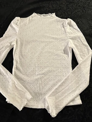 Vit långärmad blus med broderade detaljer st.M  - Säljer en vit långärmad blus med broderade hålmönster och lätt puffade axlar. Toppen har en hög hals med vågig kant och är tillverkad i mjukt bomullstyg. Perfekt för dig som gillar romantiska och stilrena plagg med fina detaljer.