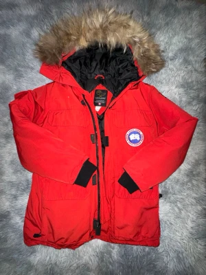 Röd Canada Goose parkajacka med päls - Säljer en röd parkajacka från Canada Goose med ikonisk patch på bröstet och stor huva med päls. Jackan har flera fickor, svarta muddar och dragkedja framtill. Perfekt för kalla vinterdagar och riktigt snygg streetstil.