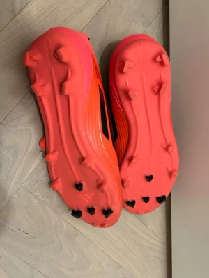 Adidas F50 orange fotbollsskor med dobbar 36,5 - Säljer ett par grymt snygga fotbollsskor från Adidas i en stark orange rosa färg med svarta detaljer och logga. Skorna har snörning, låg profil och dobbar för bra grepp på planen. Ovandelen är i syntetmaterial och insidan är svart för extra kontrast. Modell f50, som nya och endast använda 1-3 gånger. 