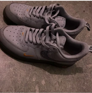 Grå Nike Air Force 1 sneakers - Säljer ett par grå Nike Air Force 1 sneakers med orange swoosh-detaljer. Skorna har klassisk snörning, perforerad tå för ventilation och låg profil. Materialet är skinn och textil, med svart sula och diskret Nike-logga på hälen. Perfekt för dig som gillar stilrena och sportiga sneakers.