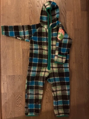 Rutig fleeceoverall från Molo - Mysig fleeceoverall från Molo i storlek 92. Overallen har ett rutigt mönster i blått, grönt, svart och vitt med gröna kantband och dragkedja. På bröstet finns en cool applikation med björntass. Perfekt för kalla dagar och lek utomhus.