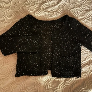 Svart glittrig kofta med paljetter - Säljer en svart glittrig kofta med paljetter över hela plagget. Koftan har lång ärm och är croppad i modellen, perfekt för att lyfta vilken outfit som helst. Materialet känns mjukt och stretchigt, och den har en öppen front utan knappar.