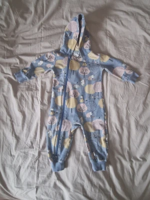 Mumin onepiece med huva - Säljer en gullig blå onepiece från Moomin med huva och dragkedja framtill. Overallen har ett mönster med Muminfigurer och träd i ljusgult, rosa och blått. Mjukt bomullsmaterial och ribbade muddar vid ärmar och ben. Perfekt för mysiga dagar hemma. Den är något tvättpåverkad i färgen.