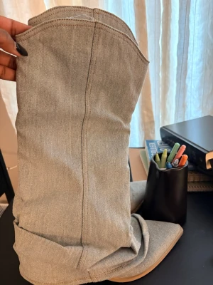 Grå denim boots med spetsig tå - Säljer ett par boots i ljusgrå denim med snyggt slouchy skaft och spetsig tå. De har en platt sula och coola sömmar som ger en unik look. Perfekt för dig som gillar statement skor med jeanskänsla.