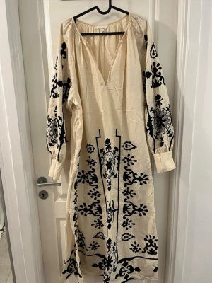 Beige långklänning/Kaftan med svart broderi H&M - Säljer en beige långklänning/kaftan från H&M i storlek XL med svart broderat mönster över hela klänningen och på ärmarna. Klänningen har v-ringad hals och långa ärmar med manschett. Materialet är bomull och designen är riktigt unik med detaljerade broderier.