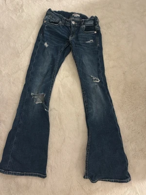 Blå bootcut jeans med slitningar - Snygga blå bootcut jeans från Gina Tricot med slitna detaljer och hål på benen. Jeansen har broderade bakfickor och justerbar midja med knappresår. Materialet är klassisk jeans i bomull.