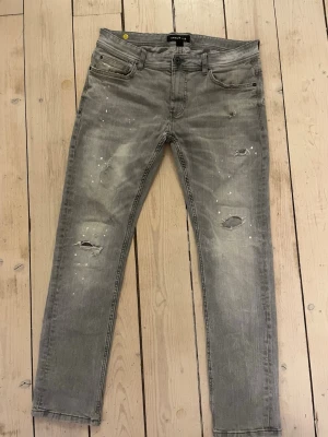 Grå jeans med slitningar från Cheap Monday - Säljer ett par grå jeans från Cheap Monday med coola slitningar och färgstänk på framsidan. Modellen är smal och har klassiska fem fickor samt knapp och dragkedja. Jeansen har en modern, lite tvättad look och är tillverkade i jeansmaterial.