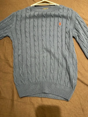 Stickad polo tröja  - Säljer en blå kabelstickad tröja från Polo Ralph Lauren med rund hals och lång ärm. Tröjan har ett orange broderat Polo-logo på bröstet och ribbade muddar vid ärmslut och nederkant. Perfekt för dig som gillar klassisk stil.