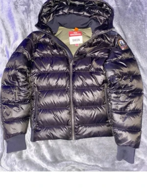 Svart pufferjacka från Parajumpers - Säljer en svart, glansig pufferjacka från Parajumpers med huva och ribbade muddar. Jackan har dragkedja framtill och en patch på ärmen. Perfekt för kalla dagar och riktigt snygg till streetwear-stilen.