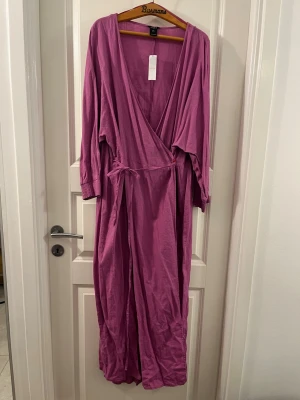 Lång lila omlottklänning från Lindex - Säljer en lång lila klänning från Lindex i storlek L. Klänningen har omlottmodell med knyt i midjan och långa ärmar. Tillverkad i ett mjukt och luftigt material som känns skönt mot huden. Perfekt för dig som gillar avslappnad och snygg stil.