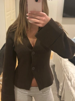 Mörkbrun kofta från Gina Tricot - Säljer en mörkbrun kofta från Gina Tricot med v-ringad hals och två stora knappar framtill. Koftan har långa ärmar med breda uppvik och en rak, något croppad passform.