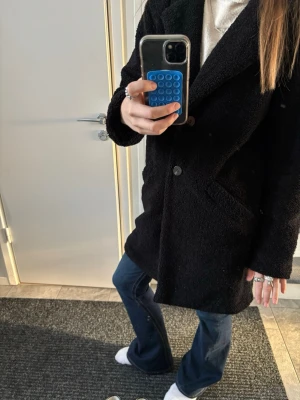 Svart teddykappa med knappar. - Mysig svart teddyjacka med två stora knappar framtill och djupa fickor. Jackan har en rak passform och är tillverkad i fluffigt teddy-material som håller dig varm. Perfekt för dig som gillar en enkel men ändå trendig look.