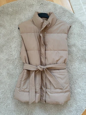 Beige dunväst med bälte L oanvänd  - Säljer en beige dunväst med hög krage och stora fickor framtill. Västen har ett quiltat mönster och knäpps med tryckknappar. Midjan markeras snyggt med ett matchande bälte som knyts. Perfekt för lager-på-lager under kyliga dagar.