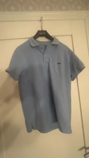 Ljusblå pikétröja från Lacoste - Säljer en klassisk ljusblå pikétröja från Lacoste med korta ärmar och den ikoniska krokodilloggan på bröstet. Tröjan har krage och knäppning framtill, perfekt för en clean och sportig stil. Inte @kt4
