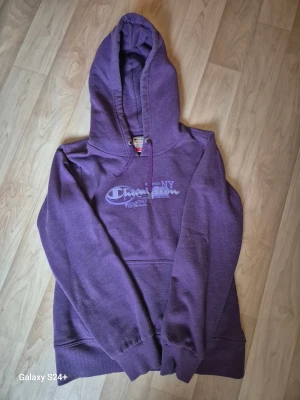 Lila hoodie från Champion, M - Säljer en snygg lila hoodie från Champion Heritage Fit i storlek M. Tröjan har huva med snörning, tryck framtill och en klassisk känguruficka. Perfekt för chill dagar och streetwear-stil. Materialet är mjuk bomull och polyesterblandning.