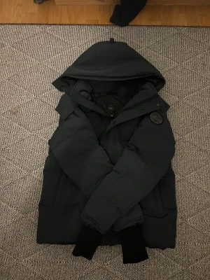 Svart pufferjacka från Canada Goose - Selling a dark green Canada Goose puffer jacket with a large hood and a stylish embroidered patch on the sleeve. The jacket features a front zipper and buttons, ribbed cuffs, and a quilted design. It has a clean, modern look and is perfect for cold days. The jacket is unused and in brand-new condition. Fits on M
