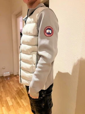 Ljusgrå Cardigan/pufferjacka från Canada Goose - Snygg ljusgrå cardigan/pufferjacka från Canada Goose med quiltad front och släta ärmar. Jackan har dragkedja, två fickor med knapp och klassisk Canada Goose patch på ärmen. Perfekt för dig som vill ha en clean och modern look.