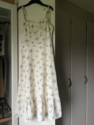 Vit blommig långklänning med volang - Superfin långklänning i vitt med små blommor i beige och grönt. Klänningen har smala axelband med knyt, volang nertill och en lätt v-ringad byst. Perfekt flowy passform och tunt och svalt tyg.😚
