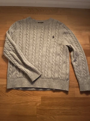 Grå kabelstickad tröja Polo Ralph Lauren s - Snygg grå kabelstickad tröja från Polo Ralph Lauren med klassisk broderad logga på bröstet. Tröjan har rund hals, ribbade muddar och är långärmad. Perfekt för dig som gillar stilrena och tidlösa plagg med premiumkänsla.