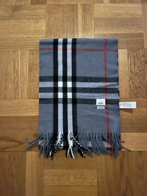 Burberry halsduk grå - Snygg halsduk från Burberry i grått med klassiskt rutmönster i svart, vitt och rött. Mjuk och lyxig känsla, tillverkad i 100% kashmir. Fransar i ändarna och tydlig Burberry-tag. Perfekt för dig som gillar stilrena accessoarer med ikonisk design.