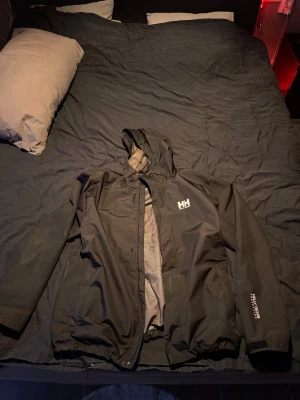 Svart Helly Hansen vindjacka 2XL - Svart vindjacka från Helly Hansen i storlek 2XL. Jackan har huva, meshfoder och HellyTech-tryck på ärmen. Utrustad med dragkedja och justerbara muddar. Perfekt för blåsiga dagar och har en clean, sportig look med HH-logga på bröstet. Köpte ny förra vintern för 2500 använde den vintern sen vart den för stor