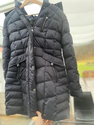 Svart dunjacka med fuskpäls från Barbara Lebek - Snygg svart dunjacka från Barbara Lebek med quiltad design och fuskpäls på huvan. Jackan har dragkedjor och knappar framtill, flera fickor med dragkedja och lång ärm. Perfekt för kalla dagar med sin långa passform och stilrena look.