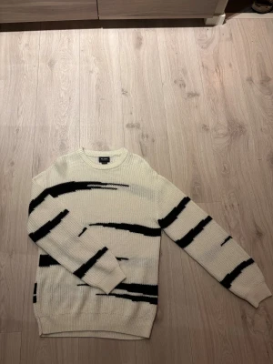 Vit stickad tröja med svarta ränder från P&B - Säljer en vit stickad tröja från pull & bear med coola svarta ränder i ett unikt mönster. Tröjan har rund hals och långa ärmar med ribbade muddar. Perfekt för dig som gillar en stilren men ändå edgy look. 