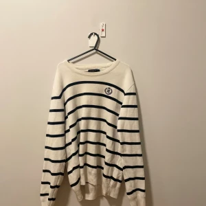 Vit randig tröja från Henri Lloyd - Snygg vit tröja från Henri Lloyd med Svarta horisontella ränder och broderad logga på bröstet. Tröjan har rund hals och långa ärmar, tillverkad i mjuk bomull som känns skön mot huden. Perfekt för dig som gillar klassisk och stilren look.