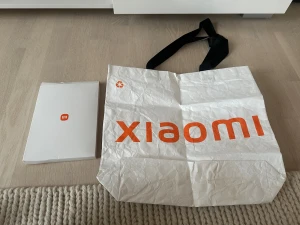 Vit shoppingväska från Xiaomi - Stor och lätt vit shoppingväska från Xiaomi med orange logga och text på båda sidor. Väskan har svarta axelband och är tillverkad i Tyvek-material som känns lite papprigt men är slitstarkt. Perfekt för att bära med sig grejer till skolan eller stan. Mått: 420mm x 380mm.