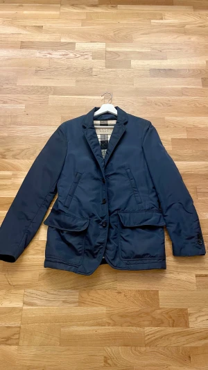 Mörkblå kavajjacka från Burberry  - Snygg mörkblå kavajjacka från Burberry med klassiskt rutigt foder i beige, svart och rött. Jackan har två stora fickor med lock, knappar framtill och quiltad insida. Materialet är polyester och jackan har en stilren, normal passform.