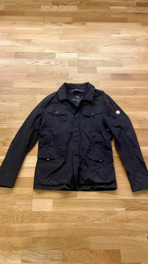 Mörkblå vindjacka från Snoot - Mörkblå vindjacka från Snoot med klassisk krage, fyra framfickor med knappar och snygg logodetalj på ärmen. Jackan är tillverkad i lätt polyester och har en stilren, normal passform. Perfekt för dig som gillar enkel och clean stil.