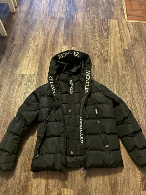 Svart Moncler pufferjacka med huva - Svart pufferjacka från Moncler med stor huva och logotyptryck på huvkanten. Jackan har två snedställda dragkedjefickor och kraftig vaddering som ger en schysst volym. Perfekt för kalla dagar och har en modern, clean look.