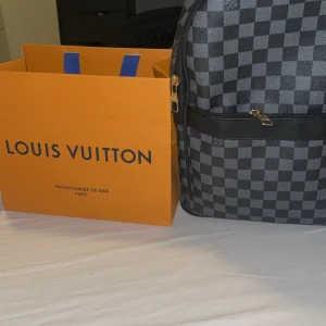 Louis Vuitton rutig ryggsäck i svart - Snygg ryggsäck från Louis Vuitton med klassiskt rutigt mönster i svart och grått. Väskan har guldfärgade dragkedjor med Louis Vuitton-logga och breda svarta axelremmar. Materialet är slitstark syntet med skinn-detaljer. Perfekt för dig som vill ha en lyxig och stilren look.