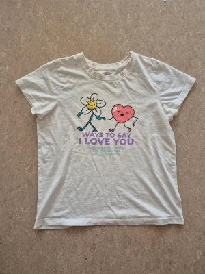 T-shirt - Vit t-shirt från Wow i bomull med färgglatt tryck framtill av en blomma och ett hjärta som håller hand. Texten 'Ways to say I love you' i lila och flera språk under. Klassisk rund hals och korta ärmar. Perfekt för dig som gillar gulliga motiv och mjuka material. Har en rosa fläck.