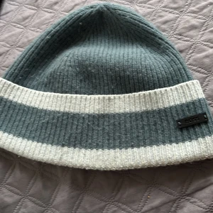 Gråvit stickad mössa från BOSS - Snygg ribbstickad mössa från BOSS i grått och vitt. Mössan har breda vita ränder nertill och en diskret BOSS-logga på sidan. Tillverkad i mjukt material som håller dig varm och passar perfekt till höst och vinter.