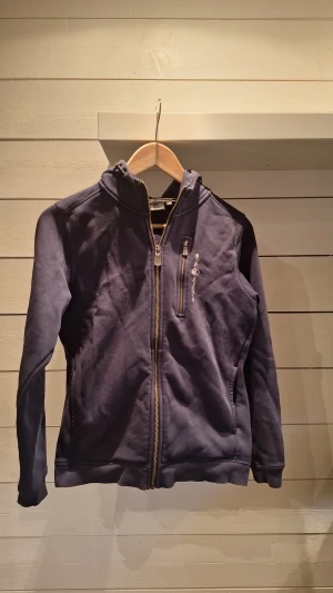 Mörkblå hoodie från Sail Racing - Snygg mörkblå hoodie från Sail Racing med dragkedja framtill och en extra zipficka på bröstet. Broderad logga på bröstet och huvan, samt justerbar dragsko. Mjuk bomullsmix som är skön att ha på sig. Perfekt för dig som gillar sportig stil.