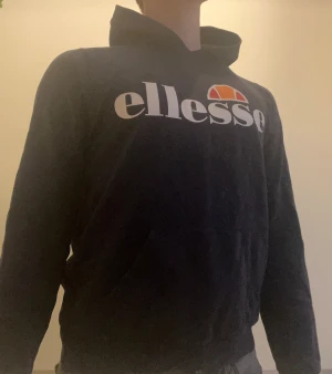 Svart Ellesse hoodie med tryck - Snygg mörk blå hoodie från Ellesse med stort vitt och orange logotryck på bröstet. Klassisk huva och magficka framtill. Perfekt för dig som gillar streetwear och vill ha en bekväm och stilren look. Mjuk bomullskänsla och långärmad modell. Hör av dig vid funderingar!