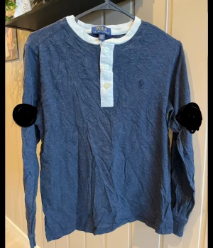 Blå långärmad tröja Polo Ralph Lauren - Snygg marinblå långärmad tröja från Polo Ralph Lauren med vit krage och knappslå. Tröjan har tre vita knappar framtill och är gjord i mjuk bomull. Perfekt för dig som gillar klassisk stil med en modern touch.