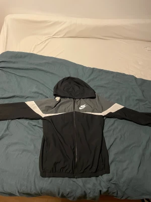 Nike svart och grå vindjacka - Nike vindjacka med huva i svart och grått med vita detaljer. Jackan har dragkedja framtill, elastiska muddar och klassisk Nike-logga på bröstet. Snygg sportig look med färgblock och långärmad design. Perfekt för dig som gillar streetwear och sport.
