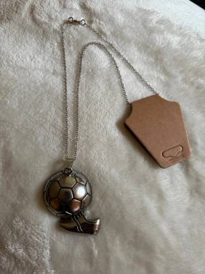 Halsband med fotbollshänge i silverfärg - Snyggt halsband med ett coolt hänge i form av en fotboll och en fotbollssko. Kedjan är tunn och silverfärgad, och hänget är detaljerat i metall. Perfekt accessoar för dig som älskar fotboll och vill visa din passion.
