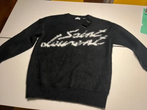 Svart Saint Laurent stickad tröja - Svart stickad tröja från Saint Laurent med vit logotyp i graffiti-stil över bröstet. Den är oanvänd och har etiketten kvar. 