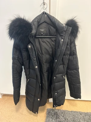 Svart pufferjacka med päls från Rockandblue - Snygg svart pufferjacka från Rockandblue med fluffig svart päls på huvan. Jackan har quiltad design, dragkedjor och knappar framtill samt flera fickor med dragkedja. Perfekt för kalla dagar och har en cool, urban vibe. Äkta päls. Köpt för 4000, använd tre vintrar. Man kan ta bort pälsen om man önskar kemtvätta den. Storlek 38 funkar som 36 också 