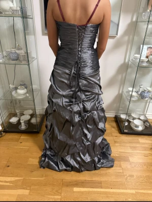 Grå långklänning med snörning - Säljer en glansig grå långklänning med smala axelband och snygg snörning i ryggen. Klänningen har draperad kjol med volanger och en dekorativ detalj med strass framtill. Perfekt för bal eller festliga tillfällen!