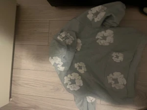 Grå hoodie med vita blommor Denim Tears - Grå hoodie från Denim Tears i storlek M med vita blommiga tryck över hela tröjan. Tröjan har huva och en stor magficka framtill. Perfekt för dig som gillar streetwear och vill sticka ut med coola detaljer.