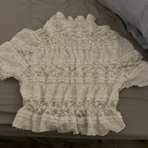 Vit spetsblus med volanger - Säljer en vit blus med spets och volanger. Blusen har korta ärmar och är dekorerad med flera lager av spets och blom detaljer. Perfekt för dig som gillar  söta toppar. Aldrig använd 