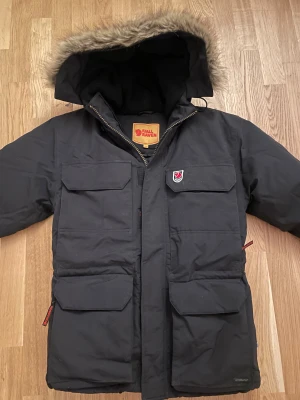 Svart parkajacka från Fjällräven XXS - Parkajacka från Fjällräven i svart med stor huva och snygg fuskpäls runt kanten. Jackan har fyra rejäla fickor framtill, dragkedja och klassisk Fjällräven-logga på bröstet. Perfekt för kalla dagar med sin långa modell och varma material.