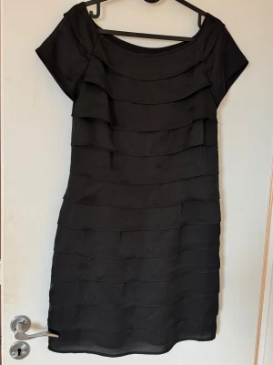 Svart kortklänning med volangdetaljer - Snygg svart kortklänning med lager på lager-volanger och rund halsringning. Klänningen har korta ärmar och en rak passform som ger en clean look. Perfekt för dig som gillar stilrena plagg med en twist. Materialet känns mjukt och följsamt.